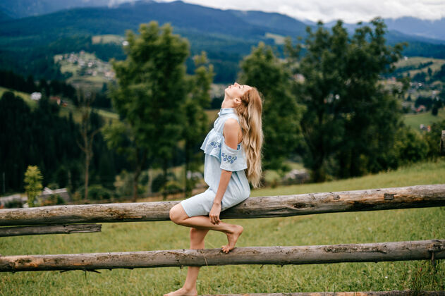 女人年轻漂亮的金发碧眼的女孩穿着蓝色的裙子坐在乡村的木栅栏上年轻服饰夏天