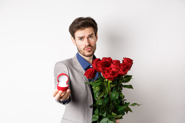 情人节穿着西装的帅哥男朋友向他求婚 手里拿着一束红玫瑰和订婚戒指 站在白色背景下 看着镜头恳求着礼物感情惊喜