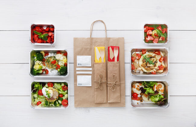 营养健康食品递送外卖食品蔬菜 肉和浆果沙拉在铝箔盒 餐具和棕色纸包装俯视图 平放在白色木材午餐蔬菜沙拉