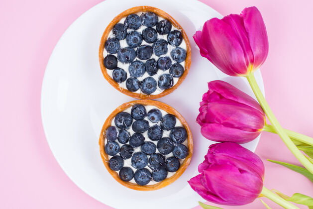酸顶部平面图俯瞰美丽明亮的花朵和两个美味的馅饼与甜蓝莓隔离粉彩柔和的表面美味奶酪惊喜