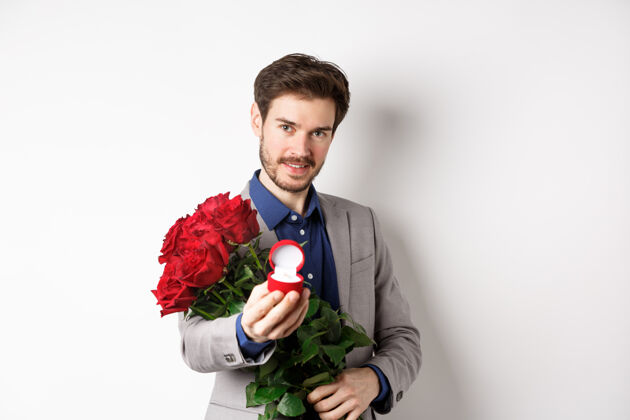 帅哥一个浪漫的男人拿着一束红玫瑰向他求婚 手里拿着订婚戒指 自信地看着镜头 穿着西装站在白色背景下丈夫男朋友男人