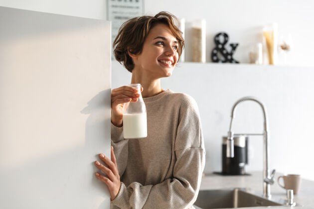 放松快乐的黑发女人喝牛奶 站在厨房里看着别处牛奶休闲冰箱