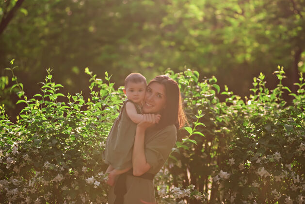春天一个年轻的女人怀里抱着一个小婴儿一位美丽的母亲和她的女儿在茉莉花丛旁的绿色公园里散步树肖像女性