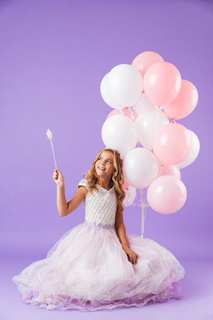 小穿着公主裙的漂亮小女孩孤立地坐在紫罗兰色的墙上 手里拿着一堆气球和一根神奇的魔杖爱裙子年轻