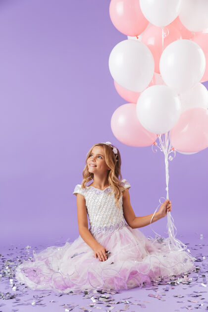 小穿着公主裙的漂亮小女孩孤立地坐在紫罗兰色的墙上 手里拿着一堆气球爱微笑快乐