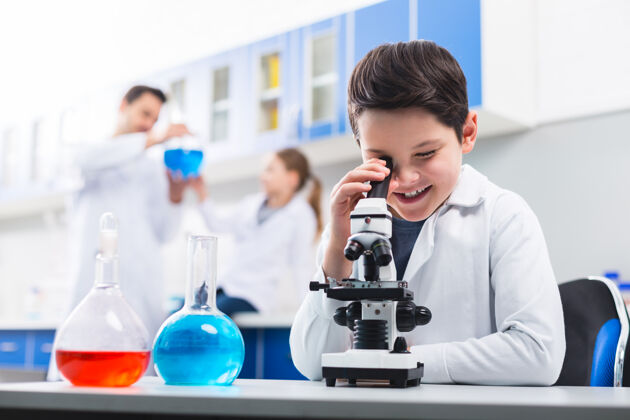 解决方案有趣的示例.创意快乐快乐的男孩一边用显微镜一边微笑着穿着实验服学习男孩年轻