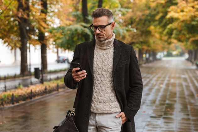 帅气30多岁的时尚男子汉穿着暖和的衣服在户外穿过秋天的公园 并使用手机的照片天气时尚成人