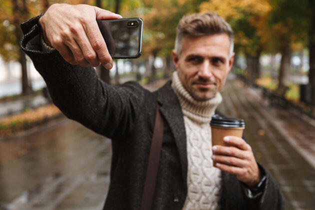 手机30多岁的笑脸男人穿着暖和的衣服在户外穿过秋园 用手机自拍的照片吸引力帅气享受