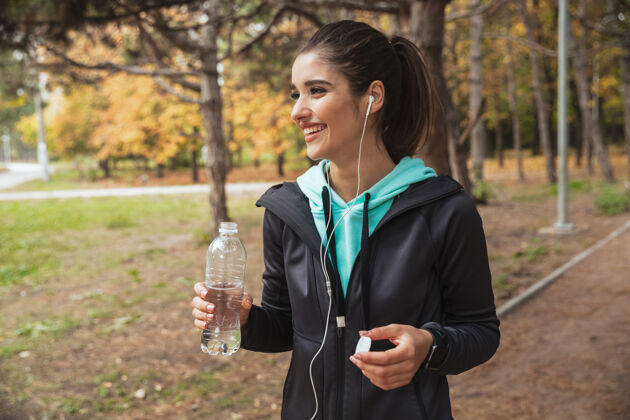 公园微笑的健身女 戴着耳机听音乐 站在公园里拿着一瓶水奉献耳机运动