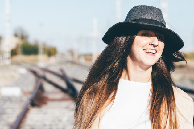 微笑在废弃的铁轨上穿着米色雨衣戴着帽子的快乐女人时尚探索行李