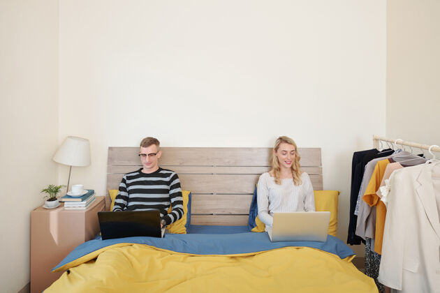 卧室快乐的年轻夫妻坐在床上 拿着笔记本电脑 工作或玩网络游戏家庭电脑女性