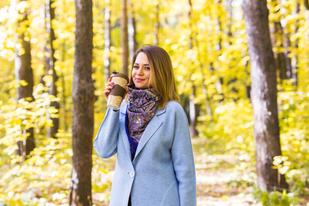 秋天穿着蓝色外套的年轻黑发女人拿着杯子站在公园里公园女性微笑