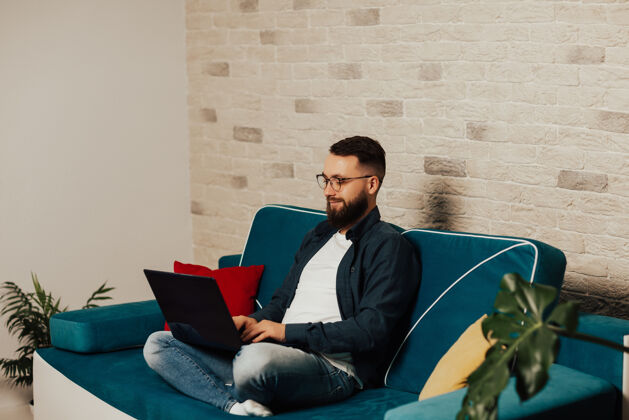 学生戴眼镜的快乐胡须男躺在客厅的蓝色沙发上 用笔记本电脑发短信微笑沟通男性