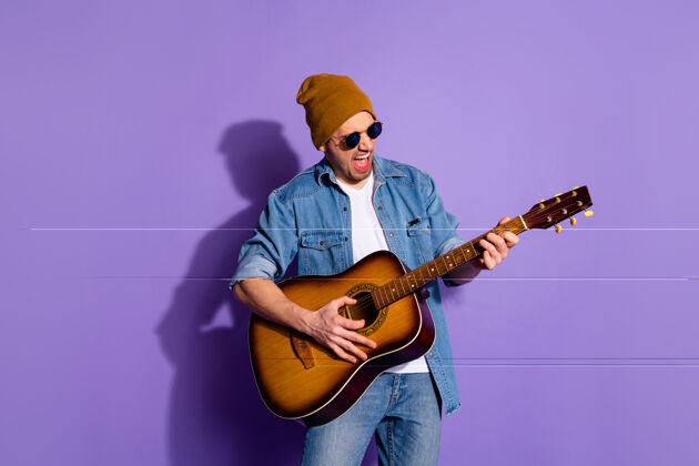 微笑照片中 开朗 粗鲁 有魅力的帅哥戴着帽子 手拿吉他 手拿乐器 戴着紫色鲜艳背景下的眼镜年轻摇滚生活方式
