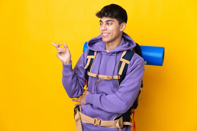 旅游者一个年轻的登山男子背着一个大背包 在孤立的黄色背景下用手指着侧面生活方式假日冒险