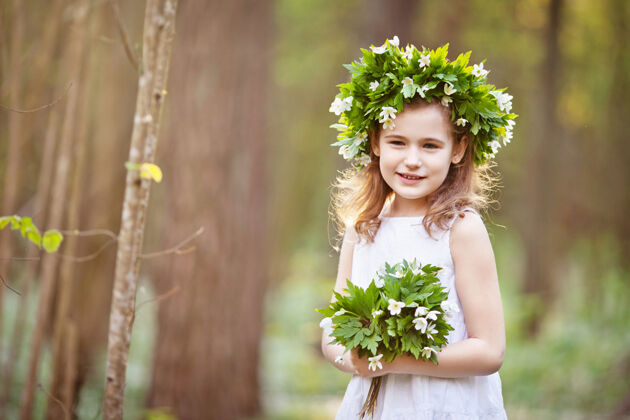 树穿着白色连衣裙的美丽小女孩在春天的树林里散步一幅美丽小女孩的肖像 头上戴着春天的花环喜爱花束童年