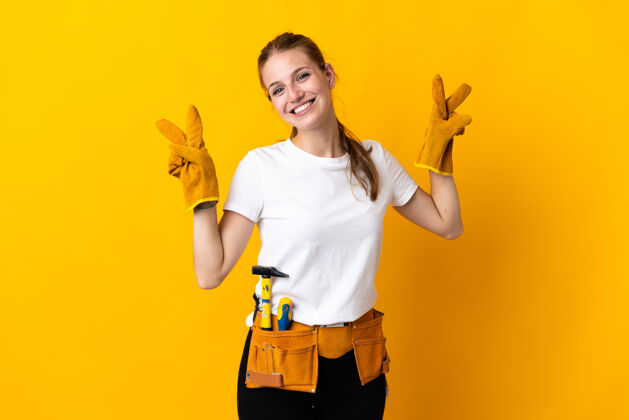 修理被隔离在黄色背景上的年轻女电工双手举着胜利的牌子工匠职业积极