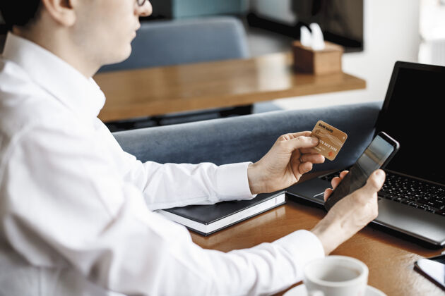 商人坐在咖啡店办公桌旁的一只男性手使用智能手机和信用卡的侧视图钱笔记本电脑电脑
