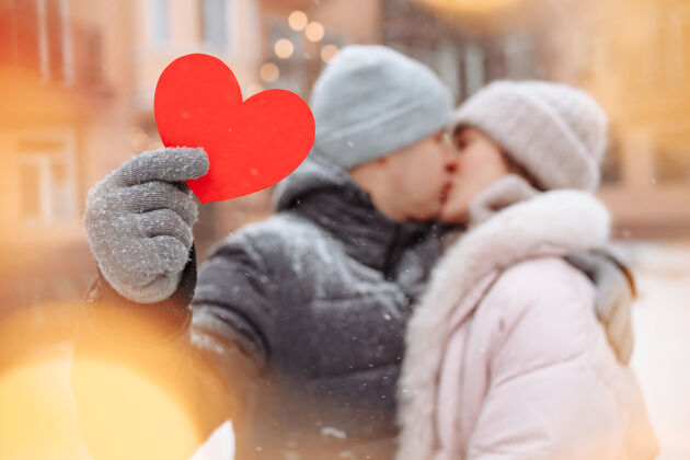 寒冷情人节的概念 相爱的情侣在冬天的雪地里亲吻拥抱帕克·杨男人拿着一个红色的纸心和他的女朋友一起庆祝情人节一对情侣在一起感觉很温暖感情享受爱