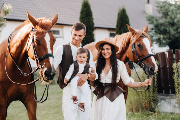妈妈一个穿着白色衣服的家庭和他们的儿子站在大自然中两匹美丽的马旁边一对时髦的夫妇带着一个孩子与马合影马连接动物