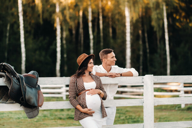 乐趣日落时分 一个戴着帽子的孕妇和她穿白色衣服的丈夫站在马厩旁边一对时髦的夫妇在大自然中等待一个孩子散步怀孕肥料