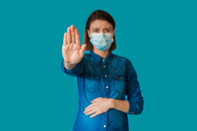 保健高加索孕妇戴着特制的口罩在蓝色背景上做停车标志的手势流感防护发烧