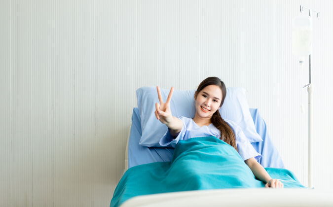 手亚洲女病人笑脸抬起两个手指在病房医院病床上与病魔搏斗孤独女人美丽