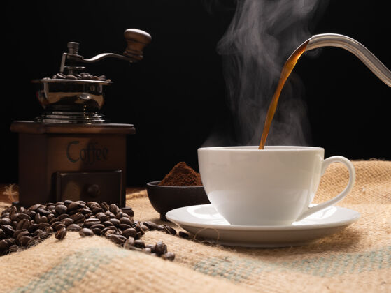 粗麻布蒸汽咖啡杯与研磨机 烤啤酒 咖啡粉和水壶在粗麻布黑森麻布在垃圾木桌子的背景桌子咖啡豆类