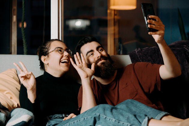 拥抱一对年轻夫妇坐在沙发上对着手机打招呼 一边微笑一边看电视女朋友男朋友社交媒体