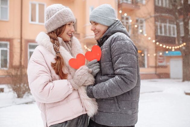 开放冬日里 一对年轻的情侣捧着红色的纸心 在外面感受着快乐和幸福爱情浪漫男人和女人在公园的雪下庆祝情人节给予男人寒冷