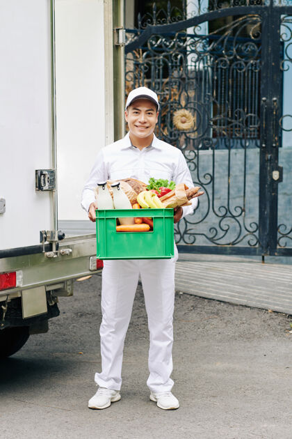 立场越南年轻帅气的信使手拿着装有牛奶瓶 新鲜面包和水果的塑料箱的画像食物商店板条箱