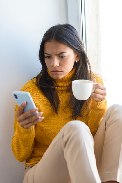 午餐特写镜头中 一个困惑的年轻女子穿着毛衣 坐在室内的窗前 端着一杯茶 用手机抱着困惑女人