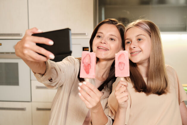 美女两位留着长发的年轻快乐的女性一边在厨房自拍 一边吃着自制的爱斯基摩草莓片冰淇淋可爱棕色头发年轻女子