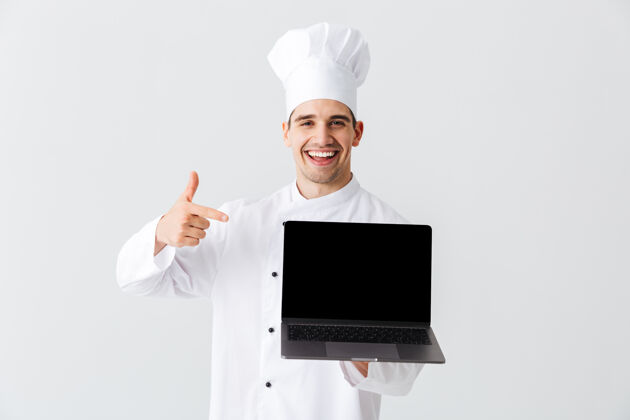 工作开朗的厨师身穿厨师制服站在白色的墙上 手里拿着银行屏幕的笔记本电脑职业男性美食家