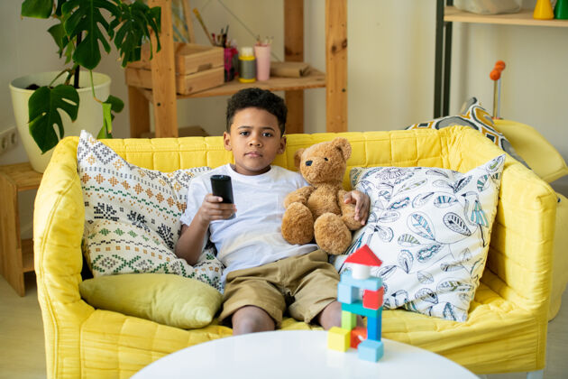 休闲服可爱的非洲学龄前儿童与遥控器和棕色泰迪熊指着你 而你躺在客厅的地板上枕头床短信