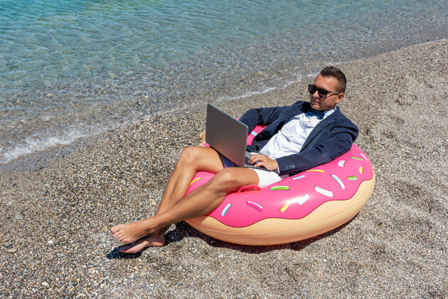 水商人在热带海滩用手提电脑做充气甜甜圈度假度假笔记本电脑