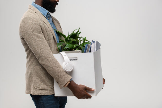 摄影棚拍摄侧视图年轻的胡须被解雇的非洲办公室经理在正装携带耳机 绿色植物和其他用品盒真人投票工作