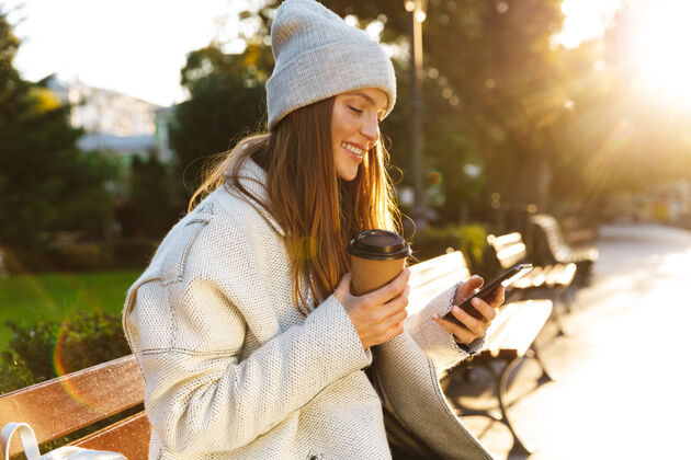 公园穿着秋衣戴秋帽的美女坐在户外的长凳上 喝着咖啡 用着手机举行年轻炎热
