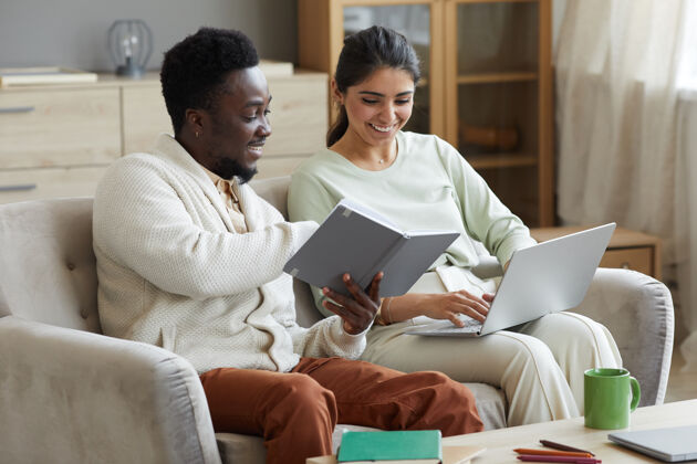 异性伴侣多民族夫妇坐在沙发上看书 用笔记本电脑上网学习互联网工作技术