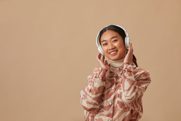 电话戴着无线耳机的亚洲年轻女子在米色墙壁上欣赏音乐的画像成人人性化音乐