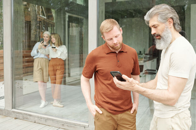 交谈大胡子男人问成年儿子智能手机应用 而他们在小屋谈论技术举行年轻人城市生活