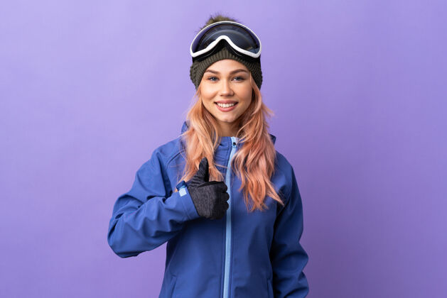 冷一个戴着滑雪板眼镜的滑雪者女孩在孤立的紫色地板上竖起大拇指表情年轻滑雪板