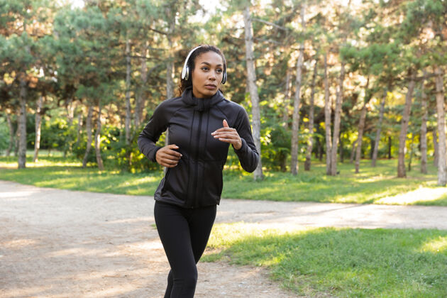 成人一个20多岁的年轻女子穿着黑色运动服 戴着耳机 一边在绿色公园里跑步健身减肥女性