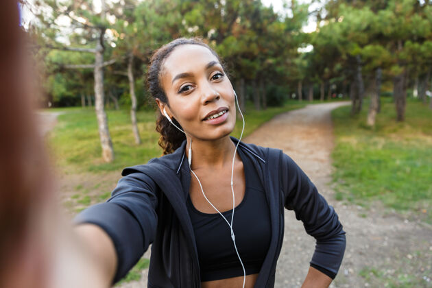 成人20多岁的美女穿着黑色运动服 戴着耳机 在绿色公园散步时用手机自拍女人火车耳机