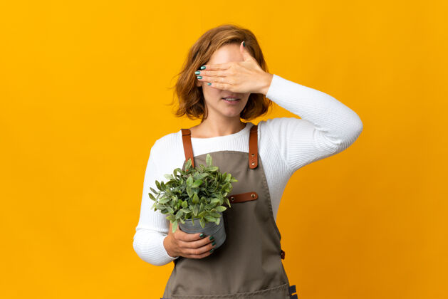年轻年轻的格鲁吉亚女子手拿一株隔离在黄色墙壁上的植物遮住眼睛不想看到什么盲人植物学格鲁吉亚