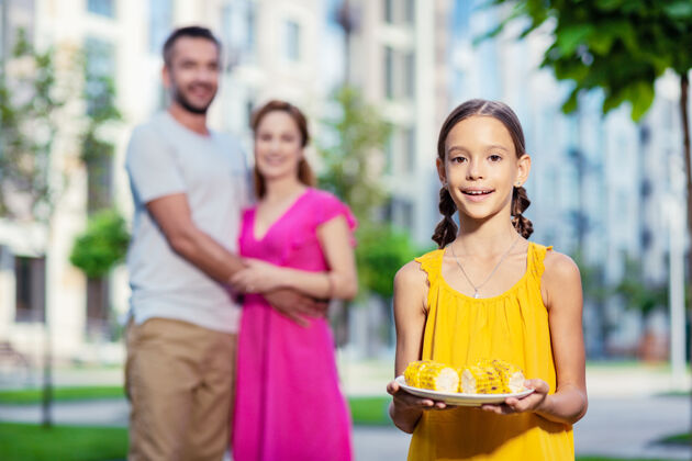 城市试试看快乐的女孩拿着一个盛着食物的盘子 她的父母站在后面人际关系美味休息