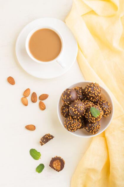 叶子巧克力焦糖球糖果杏仁和一杯咖啡在一个白色的木制表面和黄色的纺织品牛奶浪漫早餐