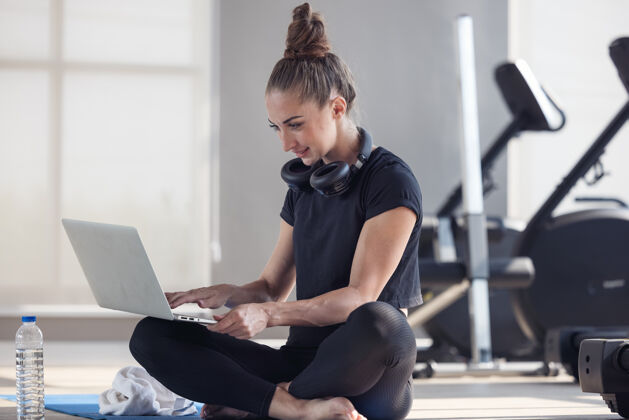 训练一个穿着运动服的运动型女人坐在地板上 手里拿着哑铃 蛋白奶昔或一瓶水 在家里的起居室里用着笔记本电脑房间.运动和娱乐理念健活方式瑜伽学习