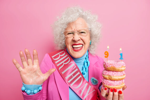 人正卷发女退休老人举着手心感到很高兴捧着一堆油炸圈饼庆祝91岁生日穿上喜庆的衣服女士乐观祖母