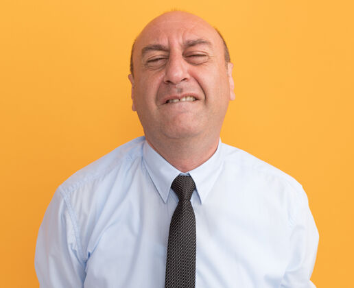 眼睛紧闭双眼紧张的中年男子穿着白色t恤 打着领带 隔离在橙色的墙上人脸姿势